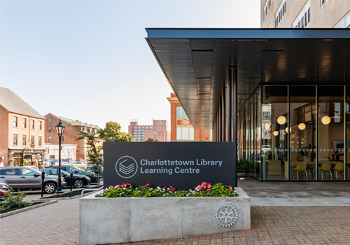 Extérieur d’une bibliothèque avec des fenêtres allant du plancher au plafond. Un panneau indiquant « Charlottetown Library Learning Centre » (Centre d’apprentissage de la bibliothèque de Charlottetown) à l’entrée.