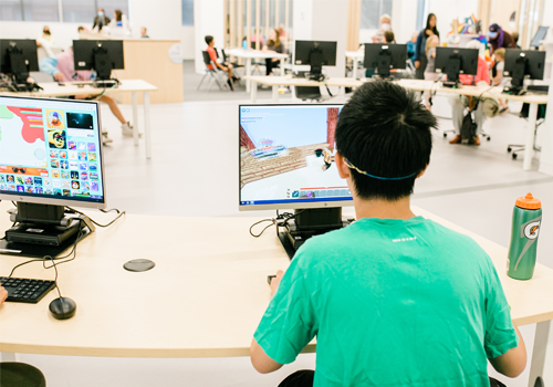 Un adolescent à un ordinateur, jouant à un jeu vidéo. D’autres ordinateurs et d’autres personnes se trouvent à l’arrière-plan d’une bibliothèque spacieuse.