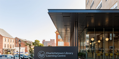 Extérieur d’une bibliothèque avec des fenêtres allant du plancher au plafond. Un panneau indiquant « Charlottetown Library Learning Centre » (Centre d’apprentissage de la bibliothèque de Charlottetown) à l’entrée.